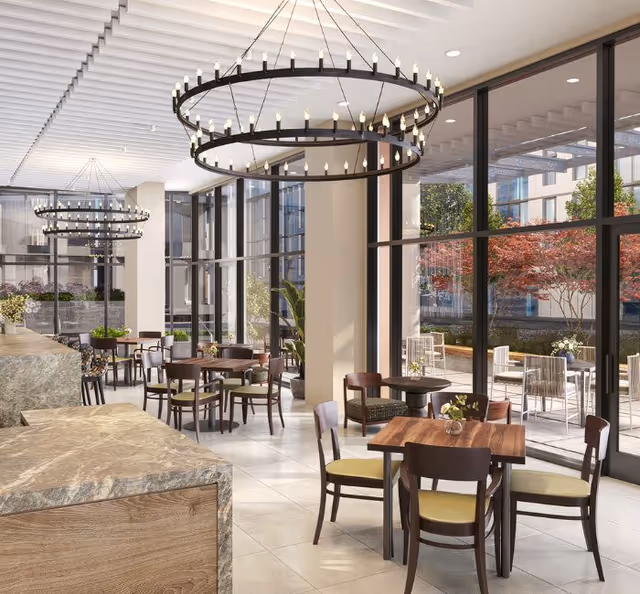 Bright dining area with multiple wooden tables and chairs featuring green cushions. Large floor-to-ceiling windows provide a view of an outdoor patio with additional seating and colorful trees. Modern circular chandeliers hang from the ceiling, and the space has a clean, contemporary design with light-colored tile flooring and a stone countertop in the foreground.