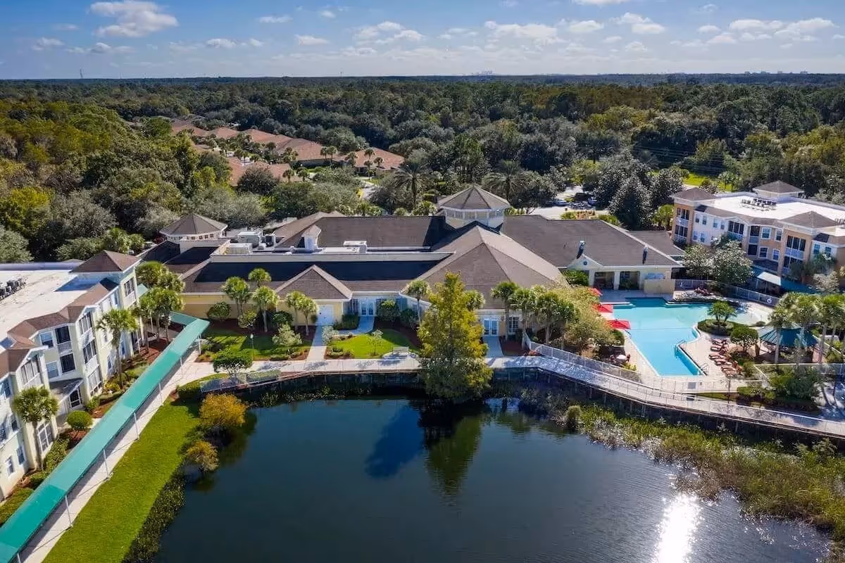 Aerial view of Aston Gardens At Tampa Bay senior living facility showing multiple buildings with beige walls and brown roofs surrounding a large pond. The facility includes a swimming pool area with lounge chairs and umbrellas, landscaped green spaces with palm trees, and a covered walkway. The background features dense green forest under a partly cloudy sky.