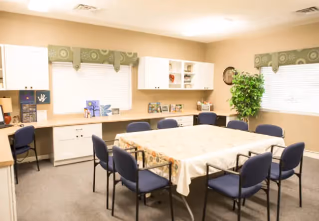 A well-lit room with a large rectangular table covered with a floral tablecloth surrounded by eight blue chairs. The room has beige walls, two windows with green patterned valances, white cabinets and shelves along the walls, and a large green potted plant in the corner. Various small decorative items and books are placed on the countertops and shelves.