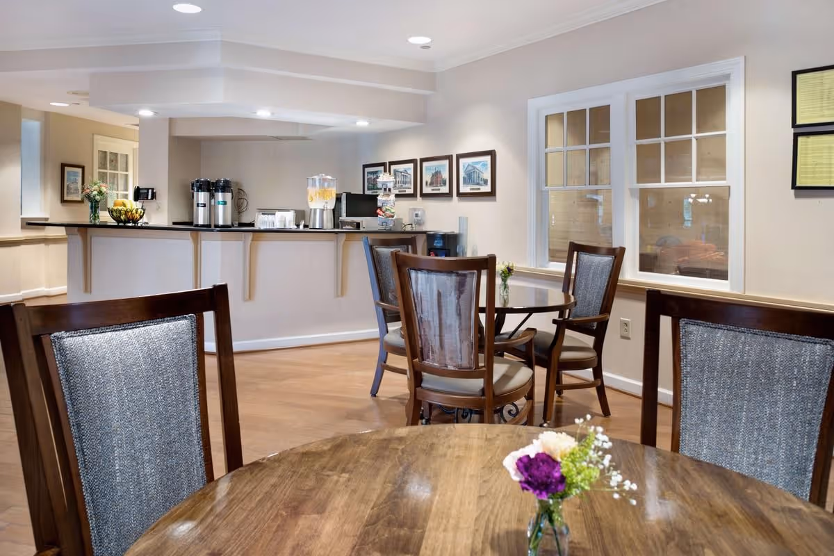 A cozy dining area in a senior living facility with wooden tables and chairs. Each table has a small vase with fresh flowers. In the background, there is a counter with beverage dispensers, cups, and a water cooler. The walls are decorated with framed pictures and windows with white trim.