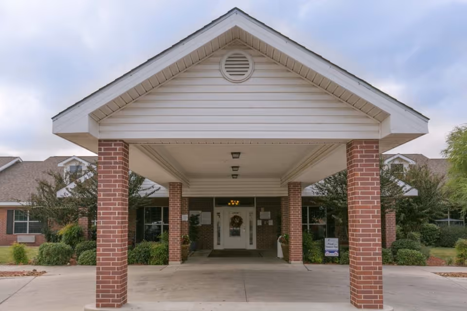 Entrance of a healthcare and rehabilitation facility with a covered driveway supported by brick pillars, surrounded by greenery and bushes under a cloudy sky.