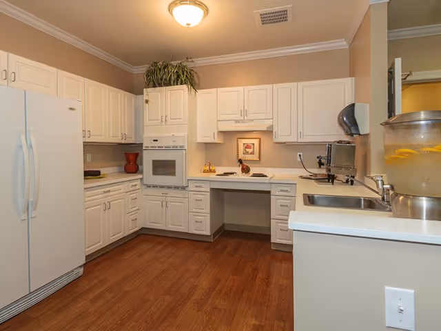 A clean, well-lit kitchen with white cabinets, a white refrigerator, built-in oven, stovetop, and a sink. The floor is wooden, and there is a water dispenser with lemon slices on the counter. A small framed picture and decorative items are placed on the countertop.