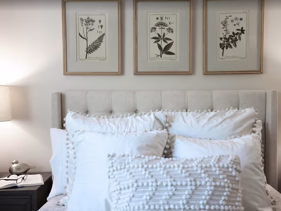 A neatly made bed with a tufted headboard and white decorative pillows beneath three framed botanical prints, with a bedside lamp and nightstand.