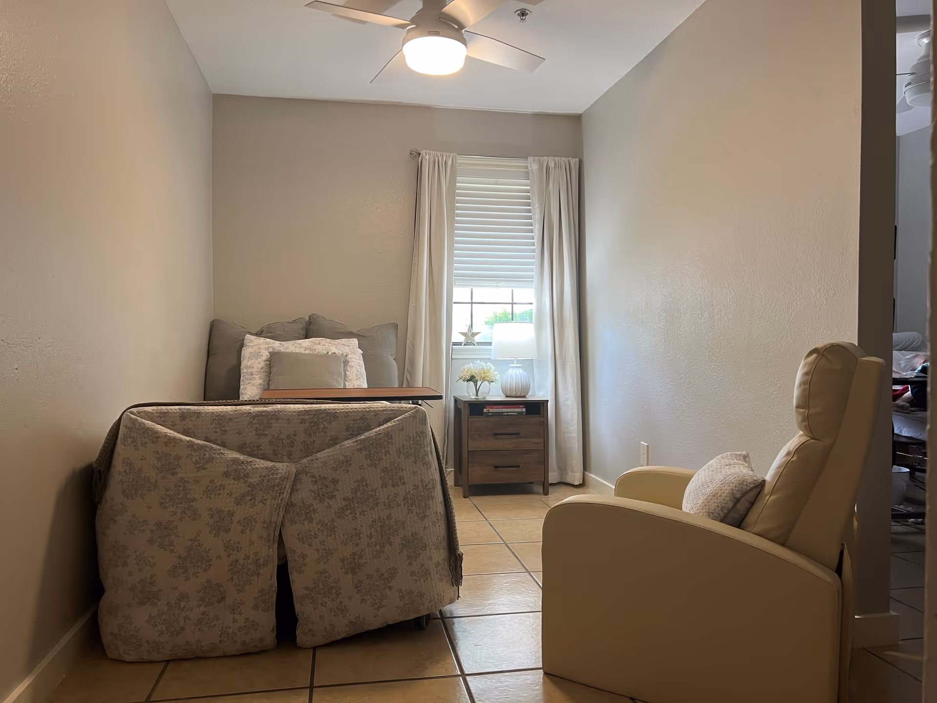 A small, cozy room with a bed covered in a floral patterned blanket and multiple pillows against the wall. Next to the bed is a wooden nightstand with a white lamp, a small plant, and a decorative star in front of a window with white curtains. A beige recliner chair with a pillow is positioned facing the bed. The room has beige walls, tiled floor, and a ceiling fan with a light.