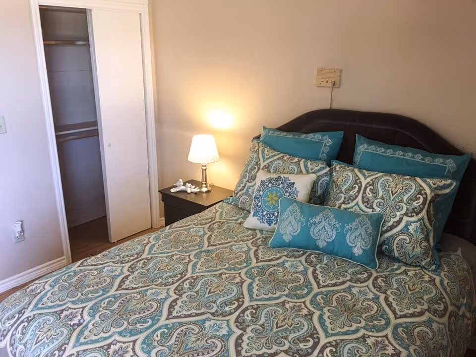 A neatly made bed with teal patterned bedding and decorative pillows beside a nightstand with a lamp and an open closet in a small bedroom.