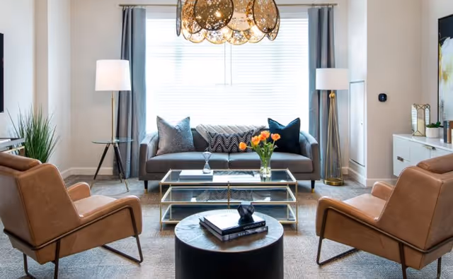 A modern living room with a gray sofa adorned with decorative pillows, two tan leather armchairs, a glass coffee table with a vase of orange tulips, a round black side table with books, two tall floor lamps, and large windows with gray curtains letting in natural light.