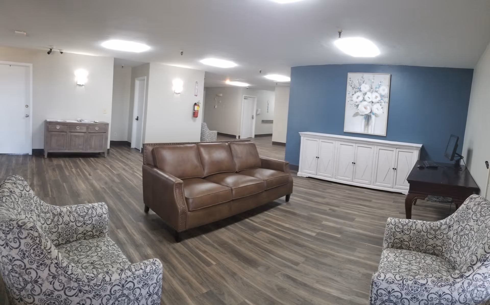 A spacious living room area with a brown leather sofa in the center, two patterned armchairs on either side, wood flooring, a white cabinet against a blue accent wall with a floral painting above it, and a dark wooden desk with a computer monitor on the right side.