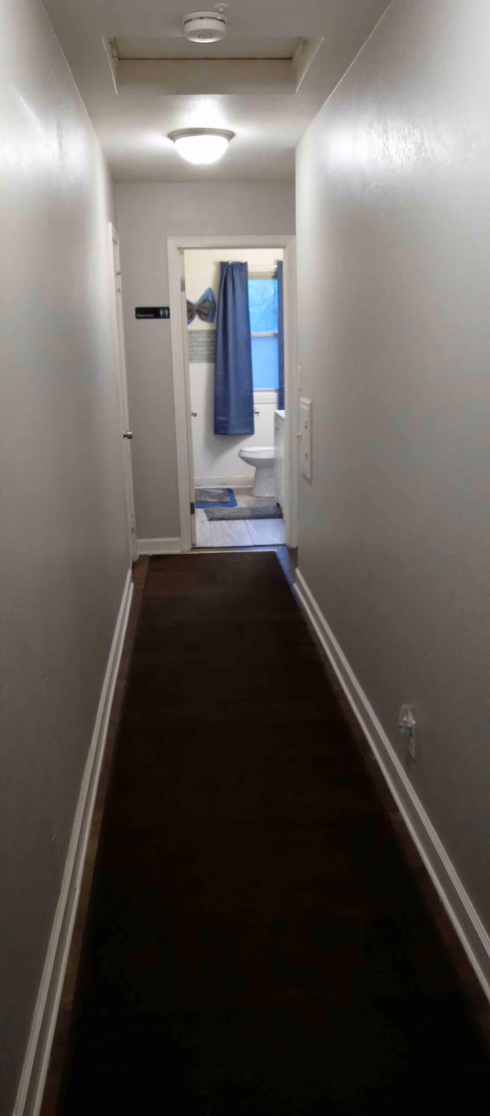 A narrow hallway with white walls and a dark carpet runner leading to a bathroom. The bathroom door is open, revealing a toilet, a blue curtain, and a window with blue blinds.