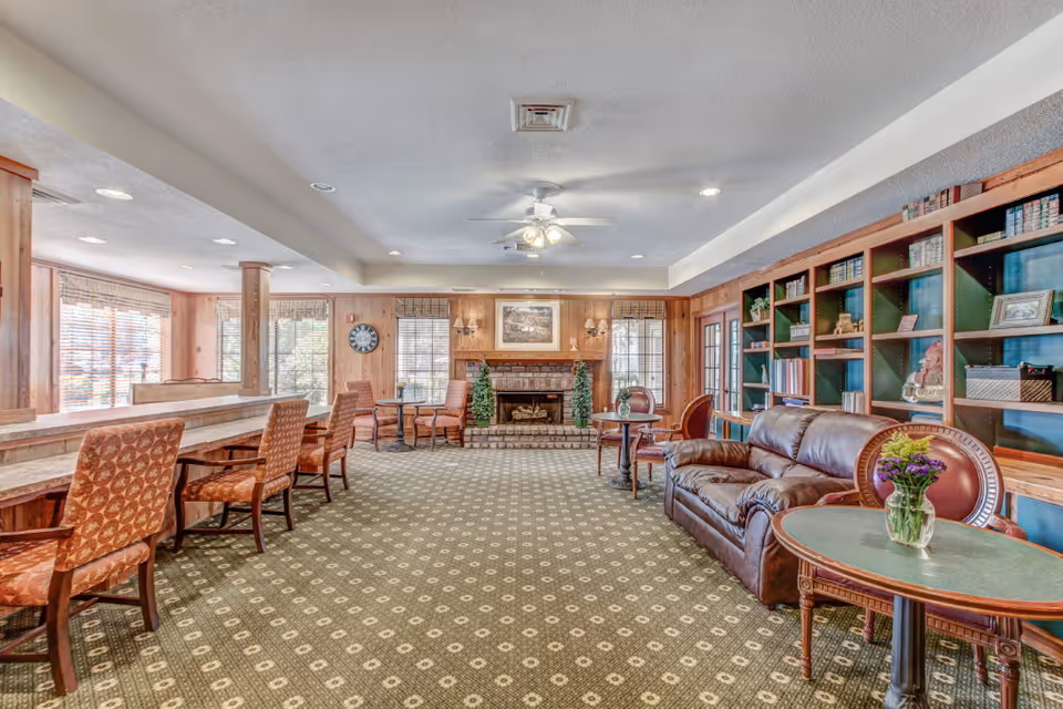 Spacious common lounge with sofas, chairs, tables, built-in bookshelves and a fireplace with large windows along one side.