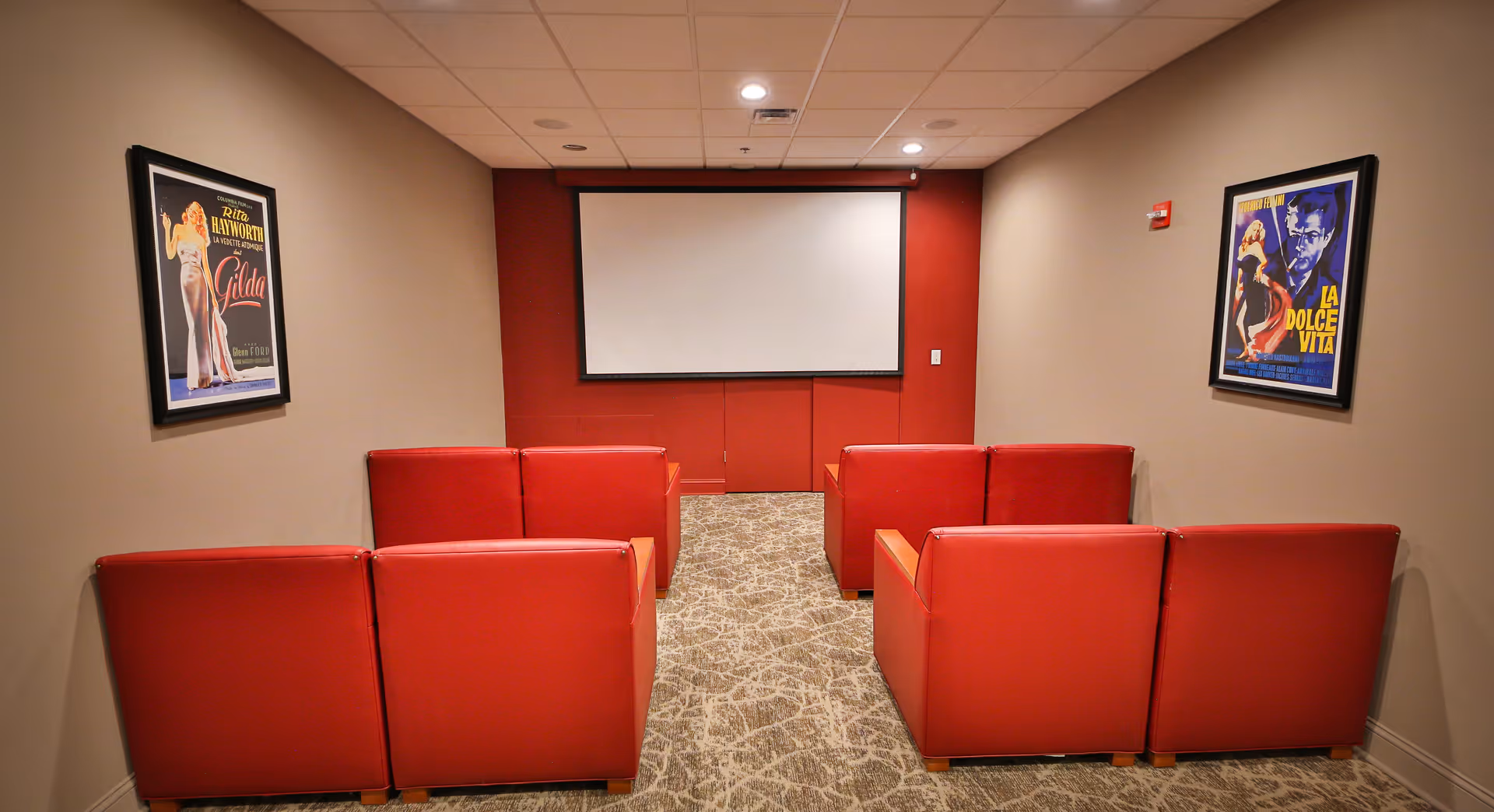 Small theater room with six red armchairs arranged in two rows facing a blank projector screen mounted on a red accent wall. The room has beige walls with framed vintage movie posters on each side and a patterned carpet floor.