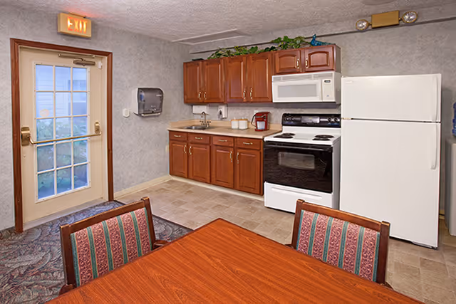 Interior view of a kitchen area in a senior living facility with wooden cabinets, a white refrigerator, stove, microwave, and a sink. There is a wooden table with chairs in the foreground and a door with a glass window and an exit sign above it on the left side.