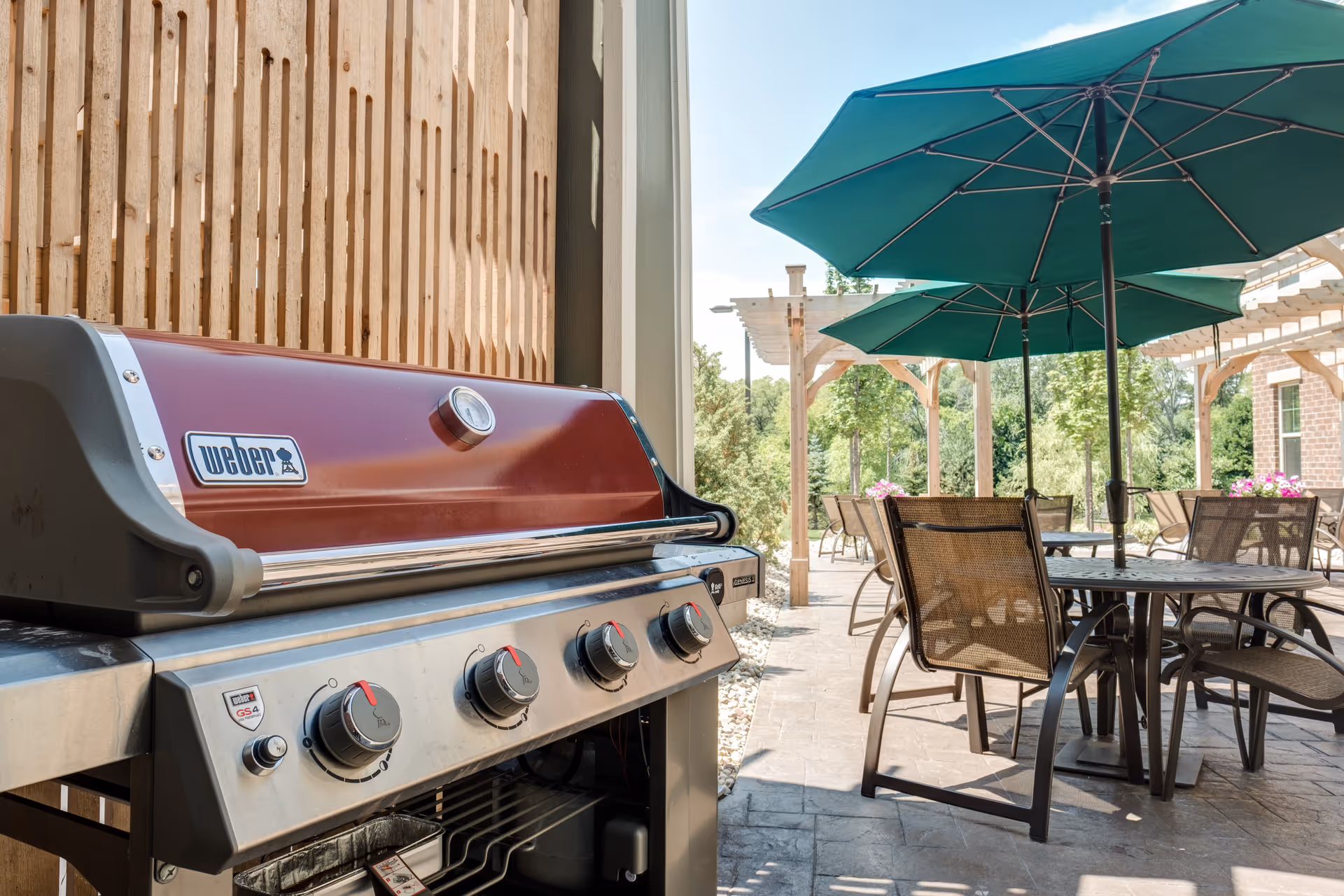 Outdoor patio area with a Weber gas grill in the foreground and several tables with chairs and green umbrellas in the background, surrounded by wooden pergolas and greenery.