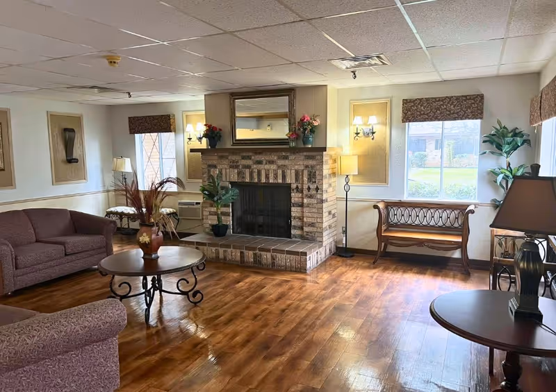 A cozy living room area with wooden flooring, a brick fireplace with a mirror and flower pots on the mantel, two large windows with patterned valances, a wooden bench, a round coffee table with a decorative vase, two upholstered sofas, and several lamps providing warm lighting.