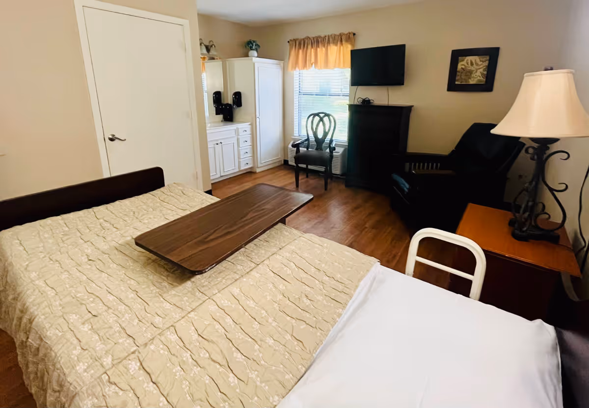 Private senior-living bedroom with a bed and overbed table, dresser, wall TV, armchair, and window.