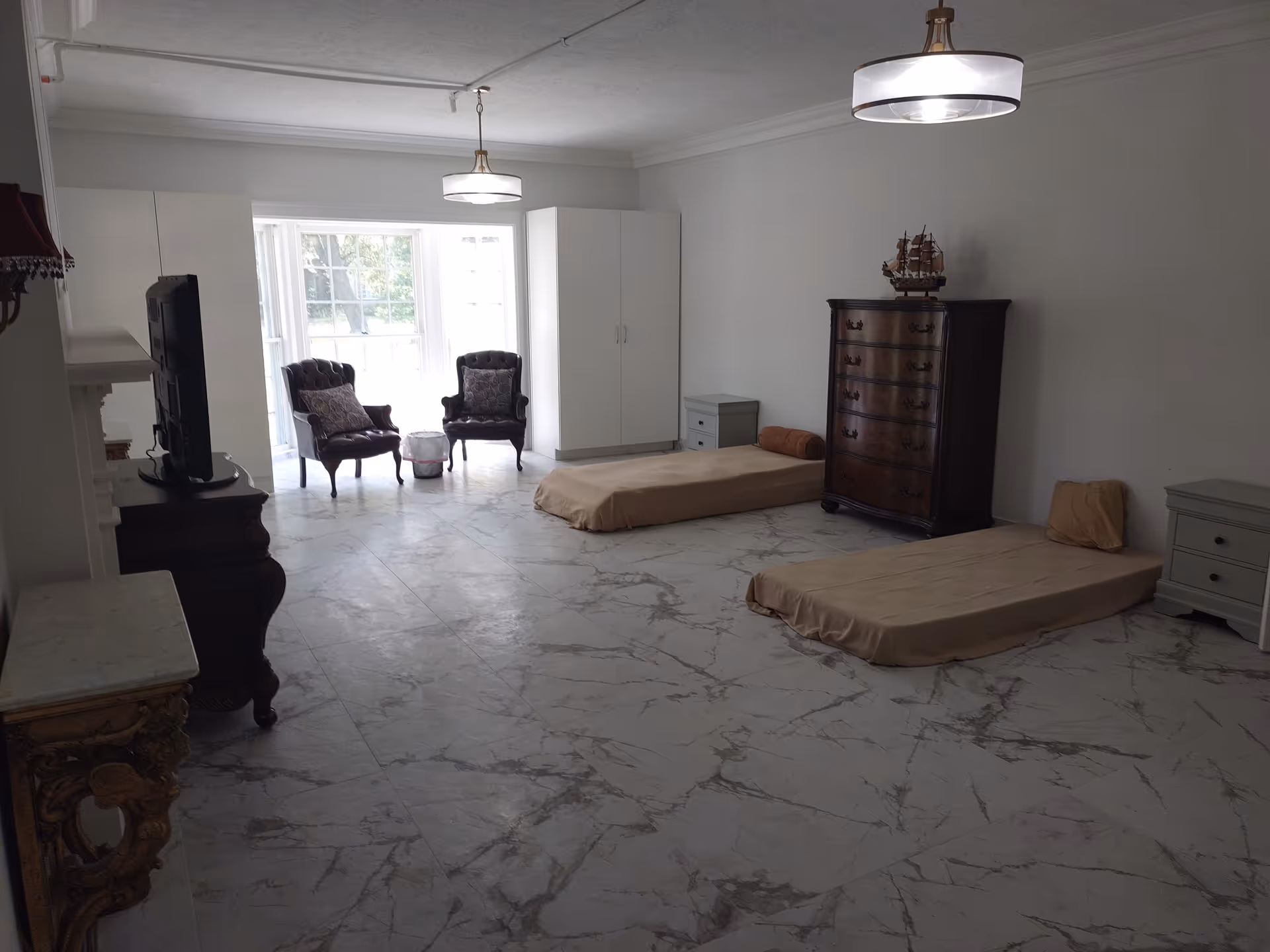 A spacious room with two low beds covered in beige sheets, each accompanied by a small nightstand. There is a dark wooden chest of drawers with a decorative ship model on top against the wall. Two upholstered armchairs with patterned cushions are placed near a large window that lets in natural light. The floor has a marble-like pattern, and two ceiling lights hang from the white ceiling.