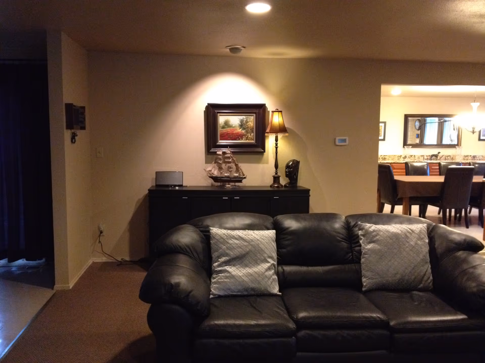 Interior living room with a black leather sofa and pillows in the foreground, a sideboard with a lamp and decorative ship against the wall, and a view into a dining area.