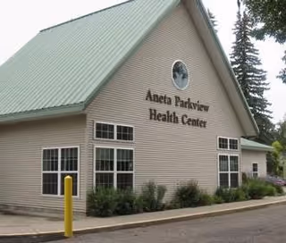 Exterior view of Aneta Parkview Health Center building with beige siding, multiple windows, and a green sloped roof. There are shrubs planted along the building and a yellow post near the sidewalk.