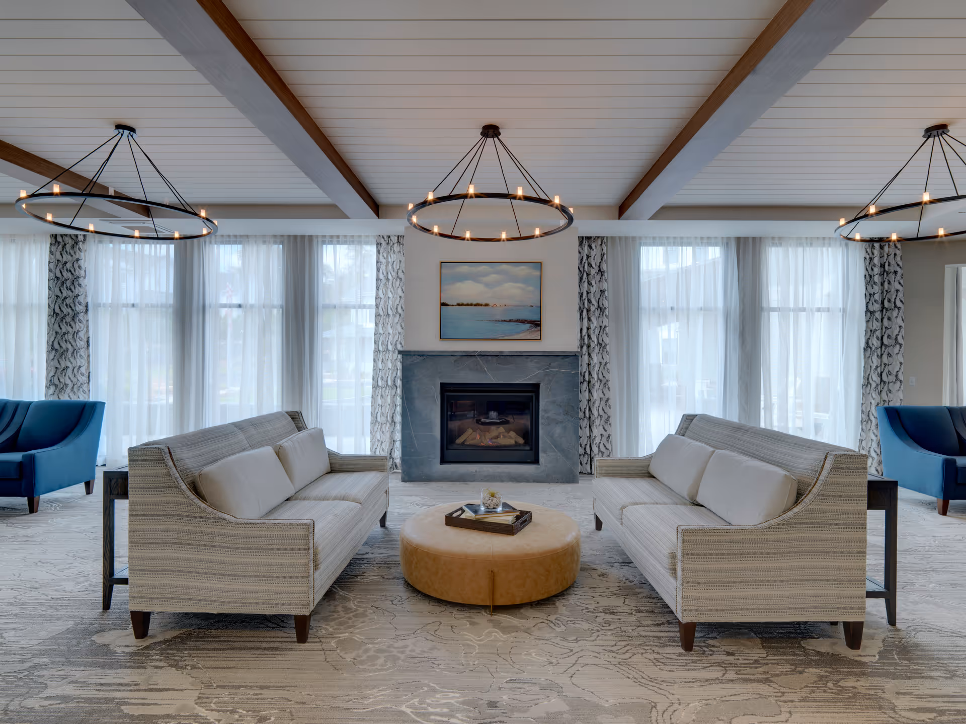 A spacious and elegant living room with two beige sofas facing each other across a round tan ottoman. Behind the sofas is a modern fireplace with a painting above it. Large windows with sheer white curtains and patterned drapes allow natural light to fill the room. The ceiling features wooden beams and three circular chandeliers with candle-like lights. Two blue armchairs are positioned near the windows on either side.