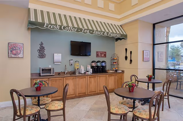 A cozy dining area with round tables and chairs featuring patterned cushions. A counter with wooden cabinets holds coffee dispensers, a toaster oven, cups, and a water dispenser. Above the counter is a green and white striped awning with the text 'Fresh Coffee - Tea - Juice & Pastries'. The walls are decorated with framed pictures and large utensils, and a flat-screen TV is mounted on the wall. Large windows allow natural light to fill the space.
