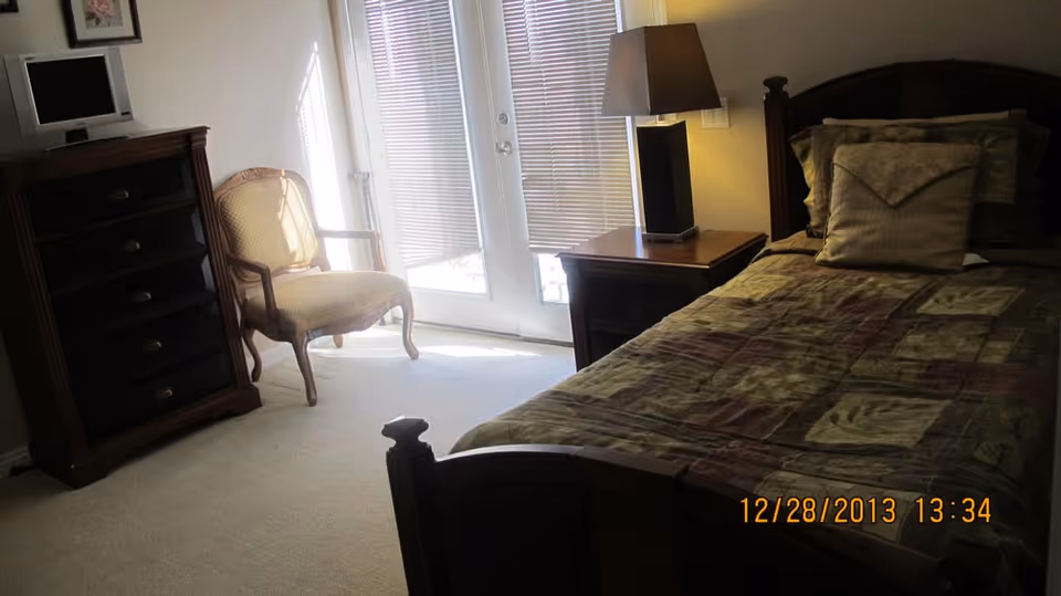 A cozy bedroom in a senior living facility with a bed, dresser, chair, and doorway.
