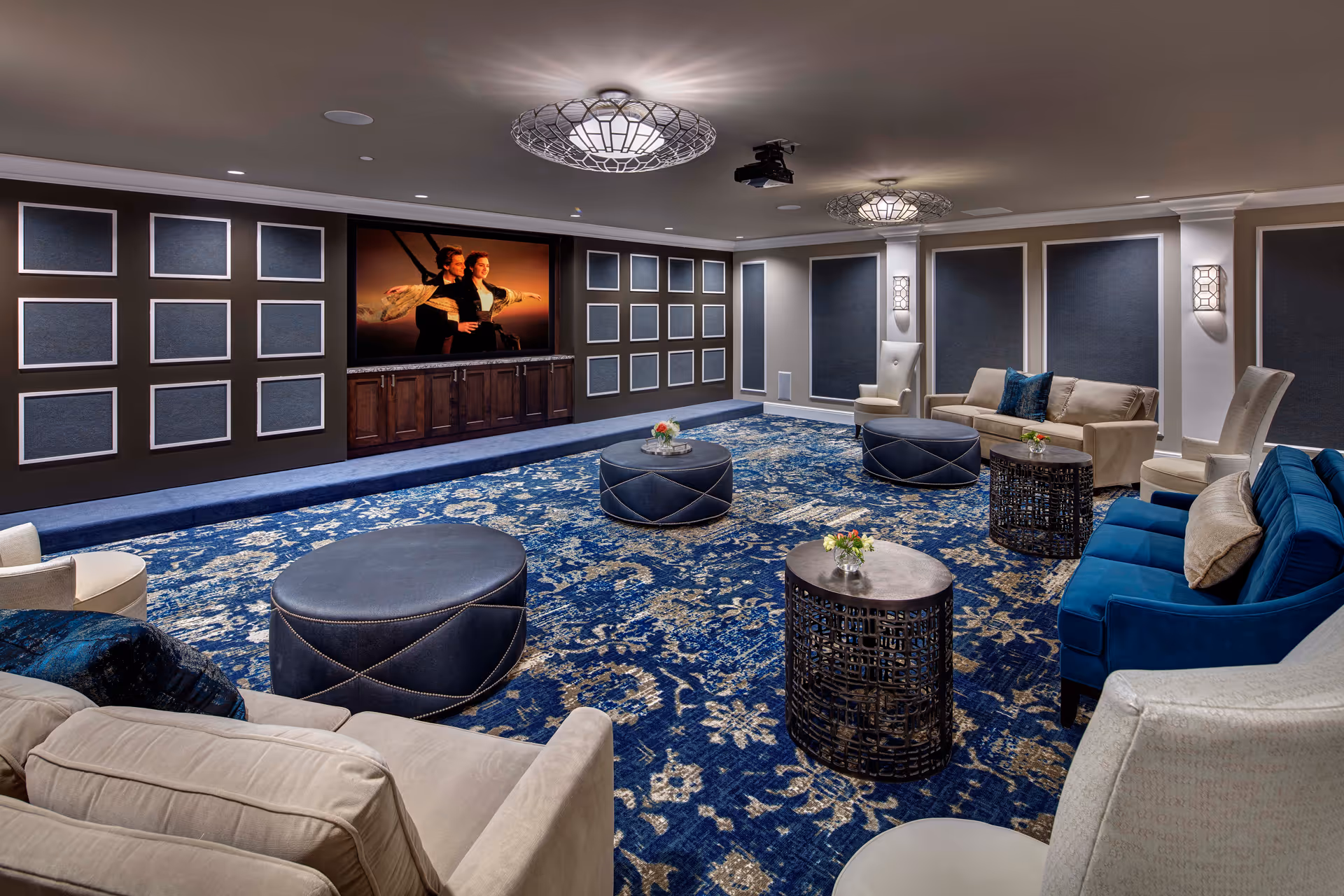 A spacious and elegant living room with a large blue patterned carpet, multiple comfortable sofas and armchairs in beige and blue tones, round ottomans, and small round tables with flower arrangements. The room features a large flat-screen TV mounted on a dark wood cabinet, decorative wall panels, and modern ceiling lights.