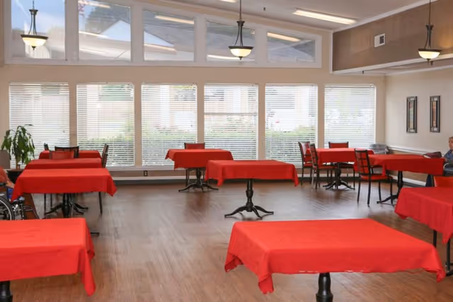 A spacious dining room with multiple tables covered in red tablecloths and black chairs. Large windows with blinds allow natural light to fill the room. The floor is wooden, and there are hanging light fixtures from the ceiling. Some framed artwork is visible on the wall.