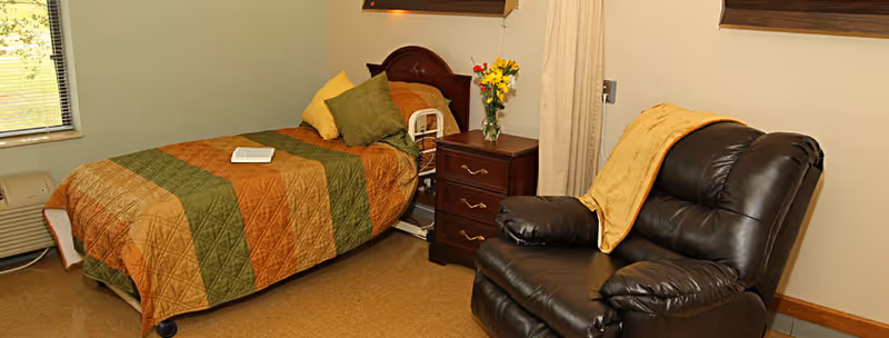 A cozy nursing home bedroom featuring a single bed with a green and brown quilt, a wooden nightstand with a vase of flowers, and a dark brown leather recliner chair with a yellow blanket draped over it. A window with blinds is partially visible on the left side.