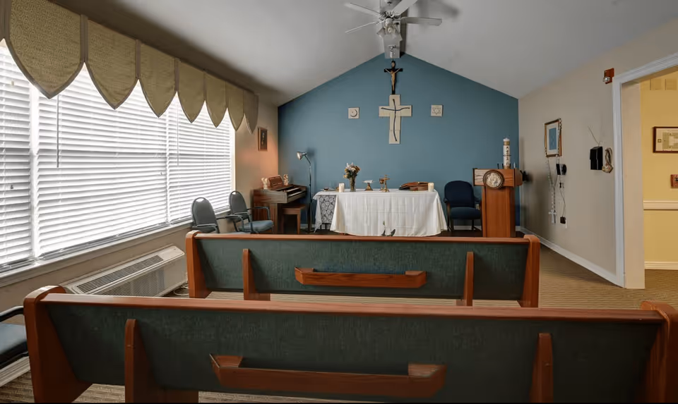 Interior view of a small chapel or prayer room with wooden pews facing an altar covered with a white cloth. The altar is set against a blue wall with religious symbols including a crucifix and a cross. There are chairs and a piano on the left side, and a lectern on the right. Large windows with blinds and decorative valances are on the left side of the room.