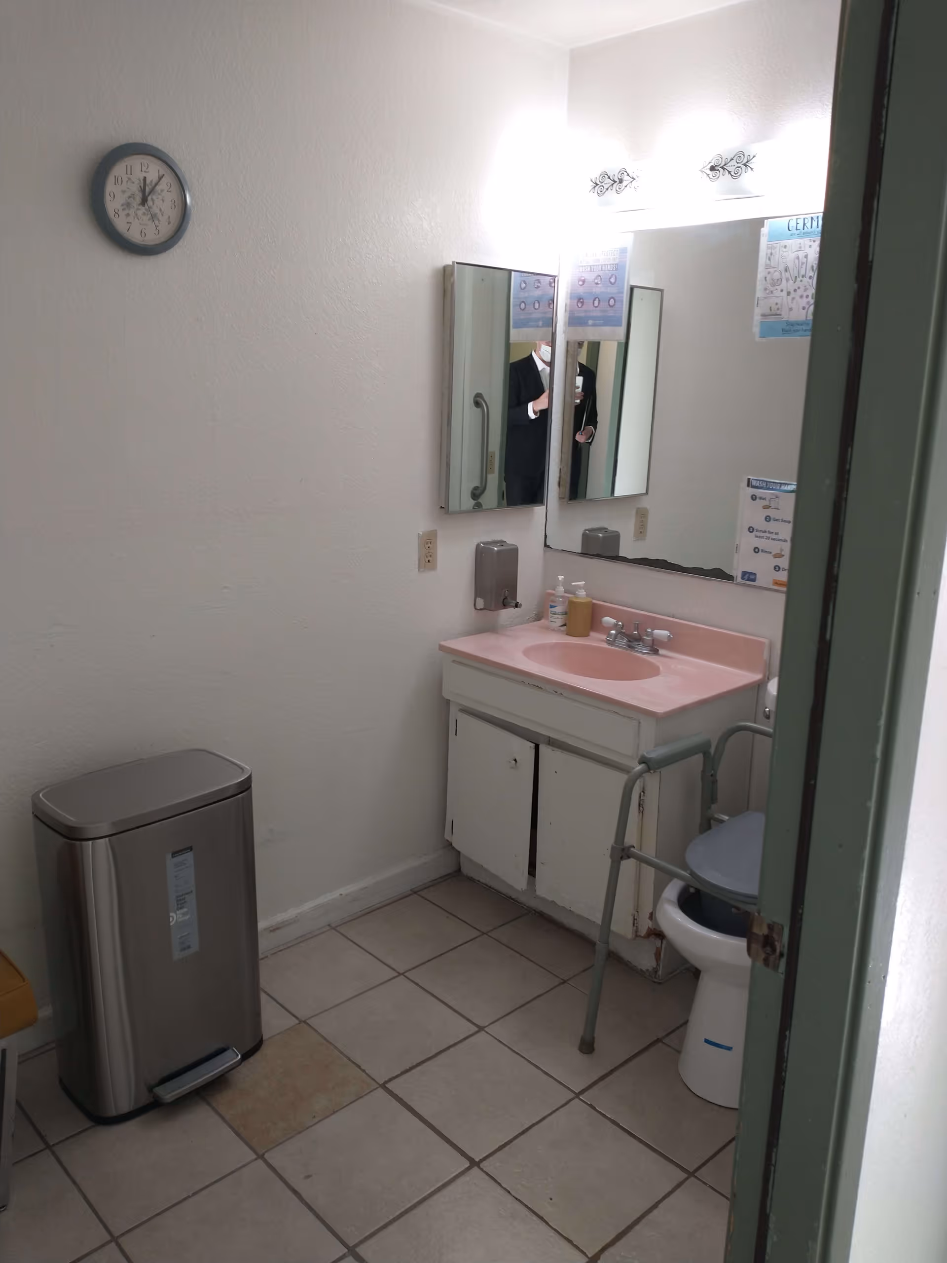 Small bathroom with a pink sink vanity, wall mirror, raised toilet seat with grab bars, and a trash can.