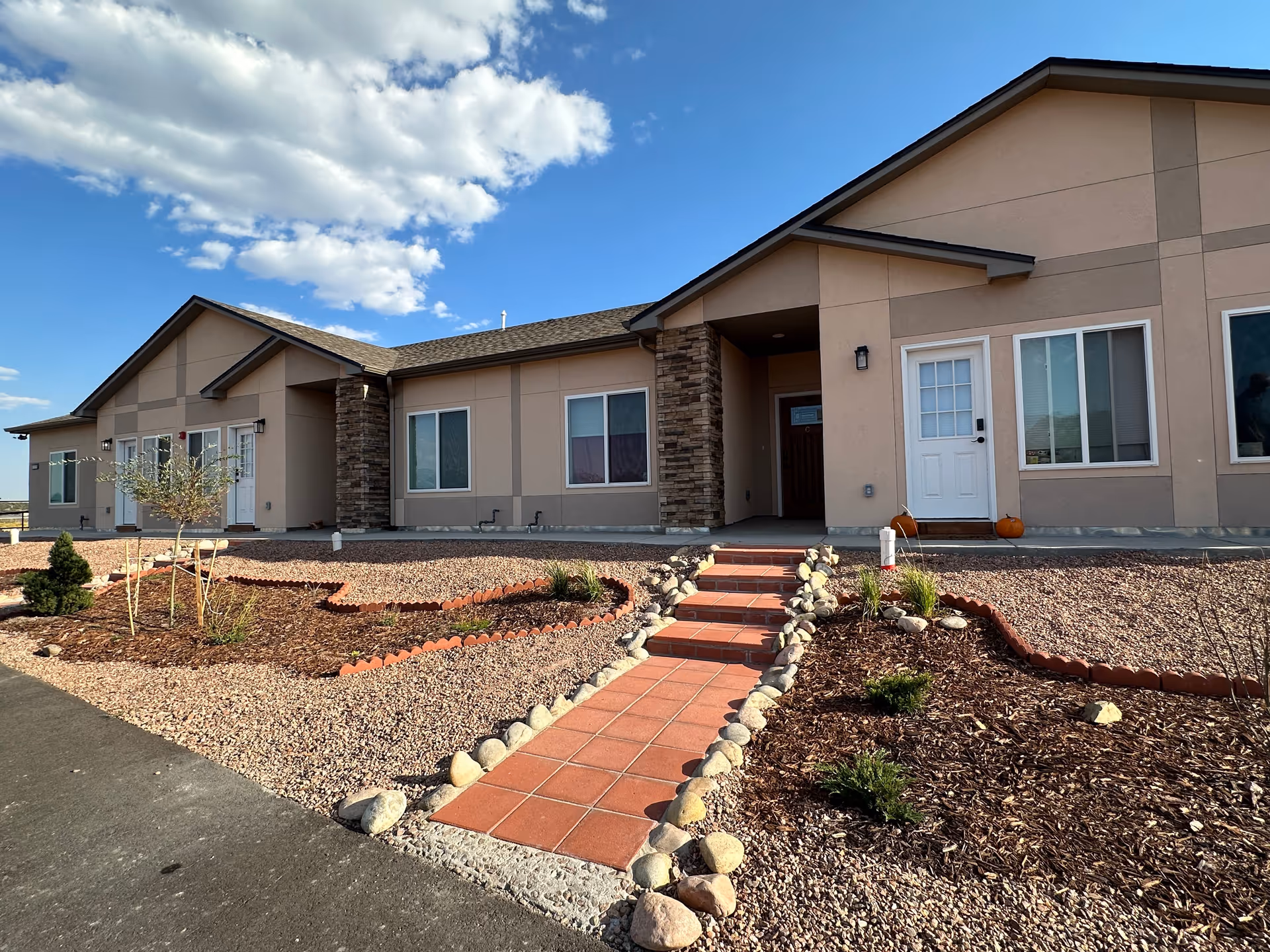 Exterior view of a single-story building with beige walls and stone accents. There is a tiled walkway with steps leading up to a white door. The landscaping includes mulch, small plants, and rocks under a partly cloudy blue sky.
