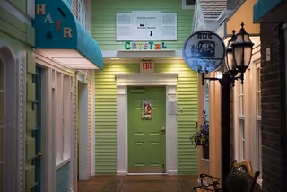 Indoor hallway designed to resemble a small town street with storefronts, including a green door at the end labeled 'Crafts', a blue awning with the word 'Hair', a hanging bus stop sign, and decorative street lamps.