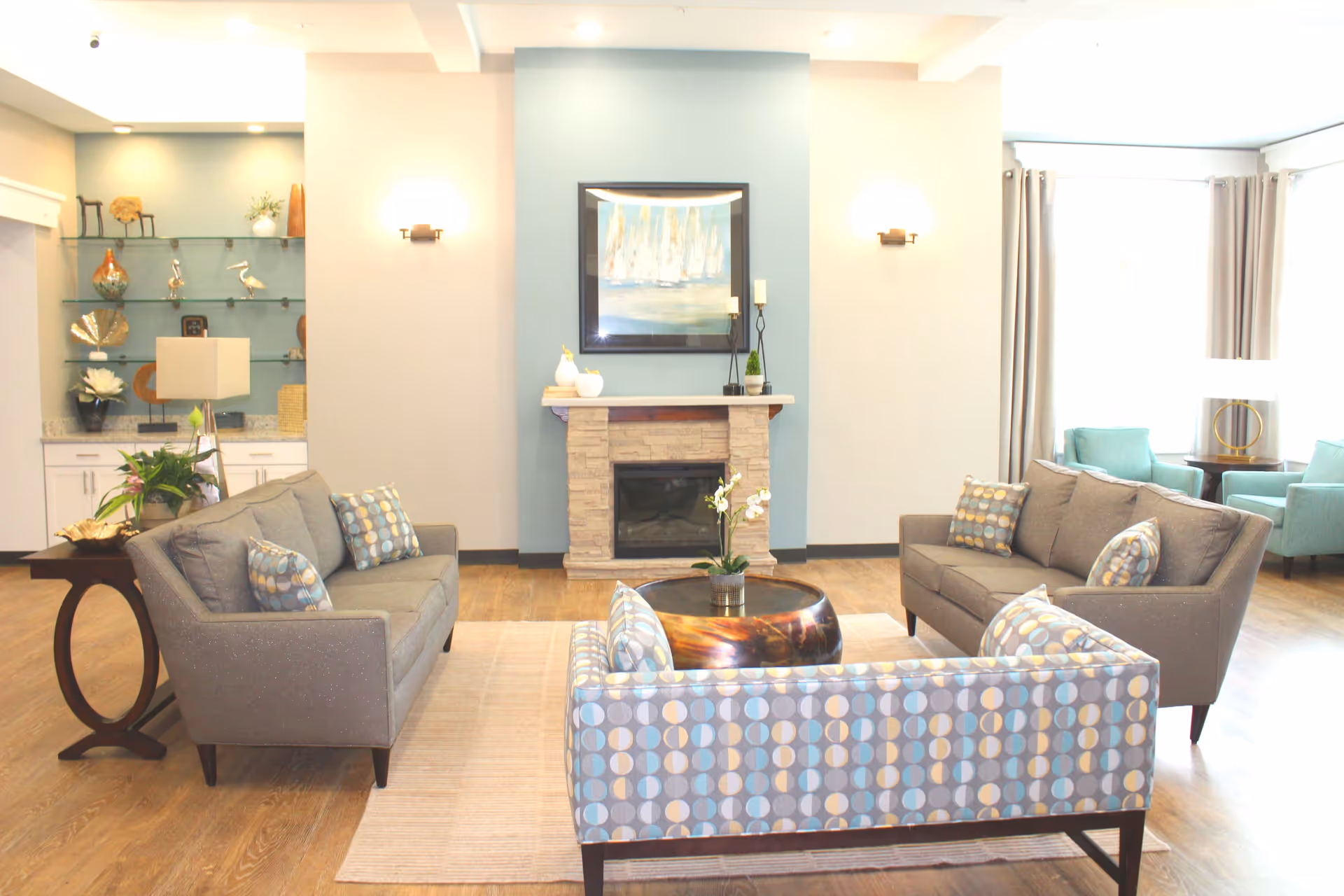 A bright and modern living room with two gray sofas and one patterned armchair arranged around a round coffee table. There is a stone fireplace with a painting above it on a blue accent wall. The room has wooden flooring, a side table with a lamp and plant, and large windows with curtains letting in natural light. Shelves with decorative items are visible in the background.