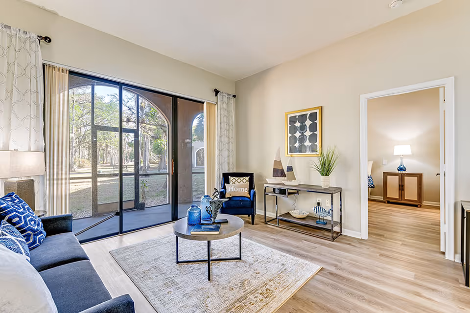A bright living room with a large glass sliding door opening to an outdoor area with trees. The room features a dark blue sofa with patterned cushions, a round coffee table with decorative items, a blue armchair with a 'Home' pillow, a console table with decorative sailboats and a plant, and a framed abstract artwork on the wall. Through an open doorway, a bedroom with a nightstand and lamp is visible.
