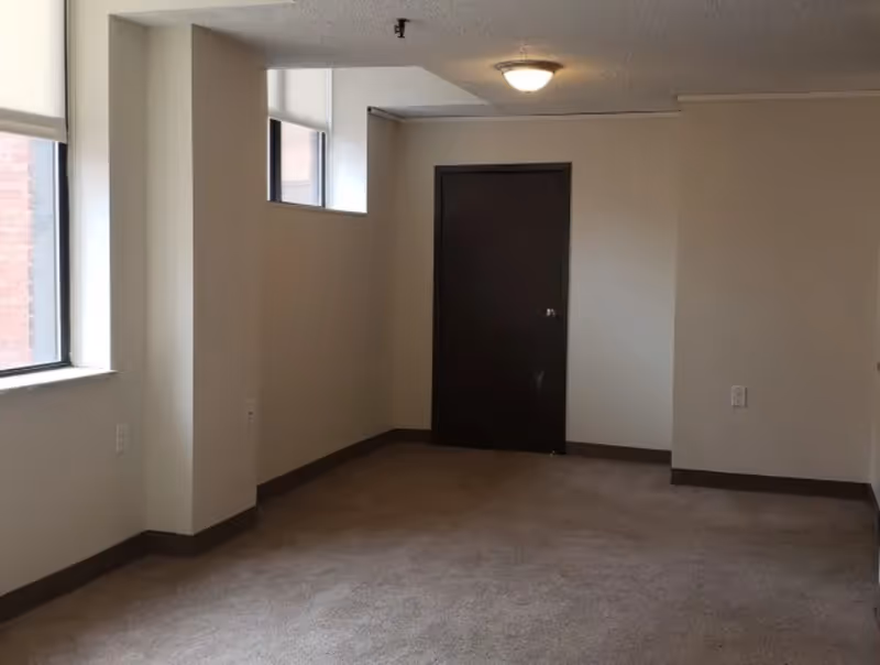Empty carpeted apartment room with a window, a small high transom window, and a closed dark door.