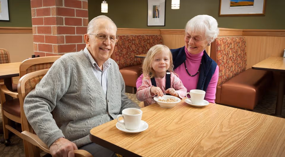 An elderly man and woman sit with a young child at a dining table with coffee cups and a bowl of cereal in a cozy dining area.