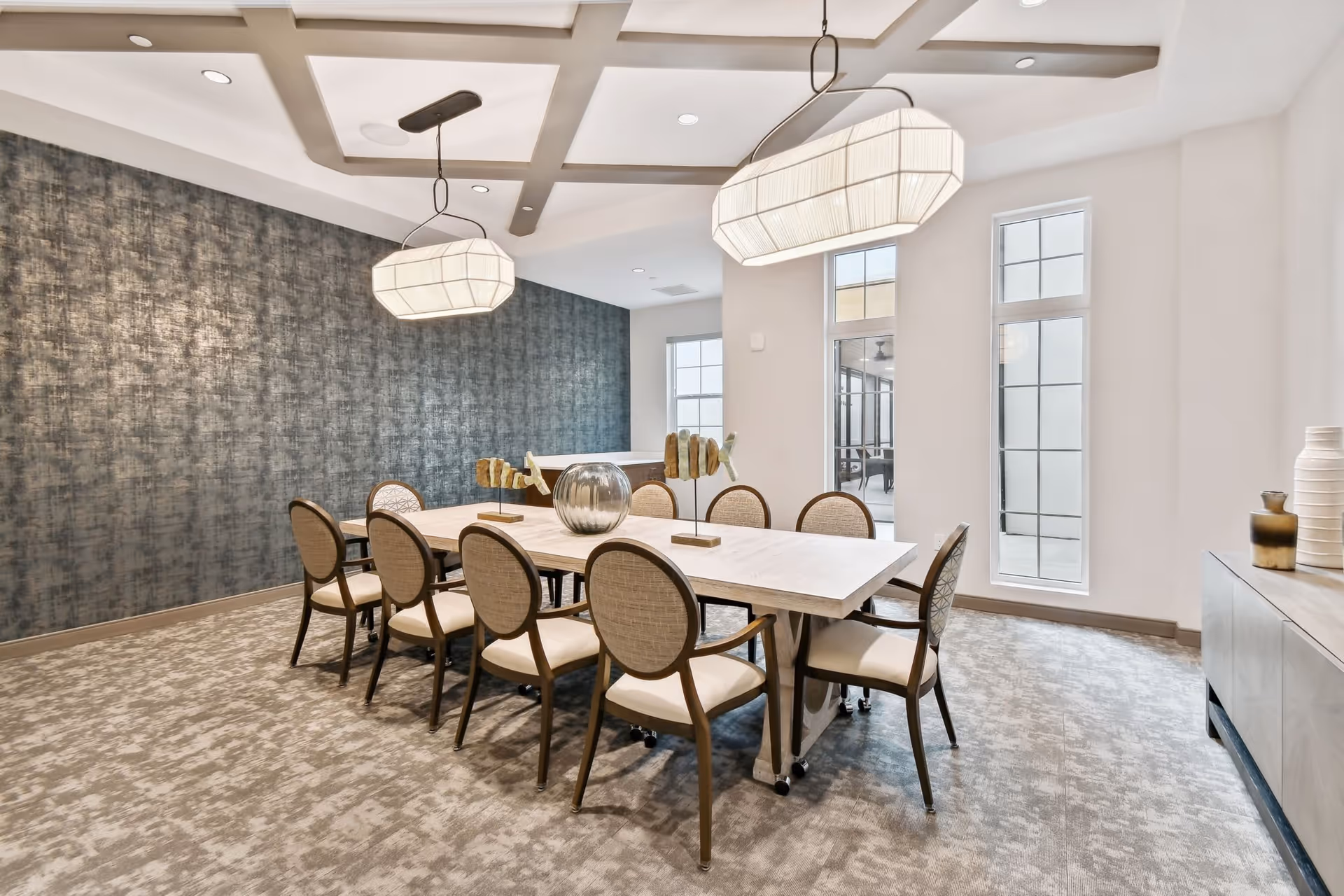 A modern dining room with a long rectangular table surrounded by eight cushioned chairs. The room features a textured gray accent wall, large windows letting in natural light, two large pendant lights hanging from a coffered ceiling, and decorative items on the table and sideboard.