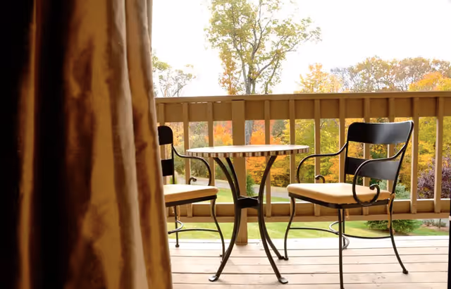 A small outdoor balcony or patio area with a round table and two metal chairs with cushions, overlooking a green lawn and trees with autumn foliage.