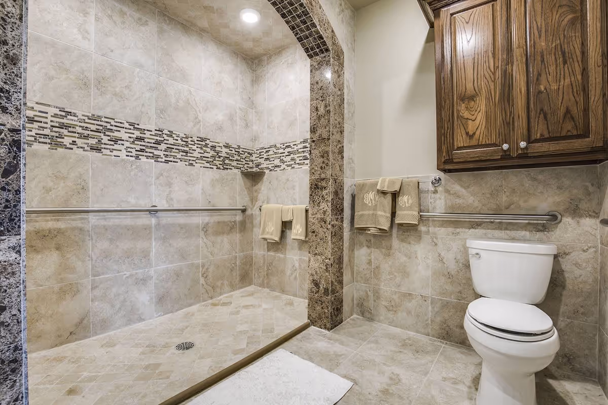 A bathroom with beige and brown tiled walls and floor. There is a walk-in shower with a tiled arch entrance and a decorative horizontal tile strip. The shower has grab bars and beige towels hanging on them. To the right, there is a white toilet with grab bars on the wall and wooden cabinets above it. A white bath mat is placed on the floor outside the shower.