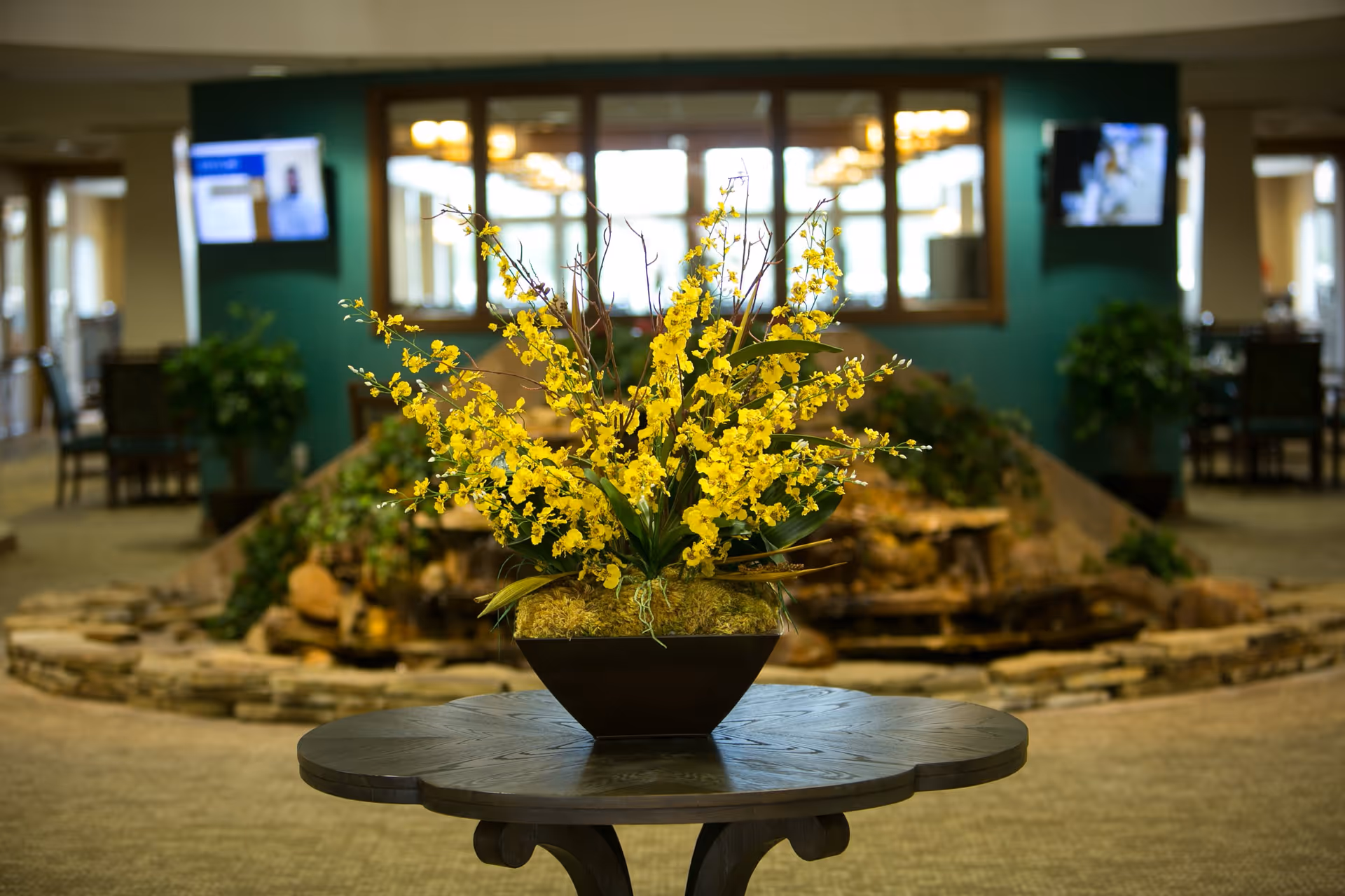 A decorative indoor arrangement featuring a vase with yellow flowers placed on a round wooden table. In the background, there is a stone water feature with greenery, two wall-mounted televisions, and a large mirror reflecting ceiling lights in a spacious, carpeted room.