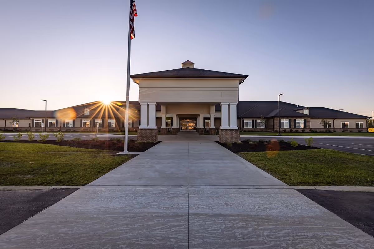 Front exterior view of a senior living facility building with a covered entrance, an American flag on a flagpole, landscaped grass and shrubs, and the sun setting in the background.