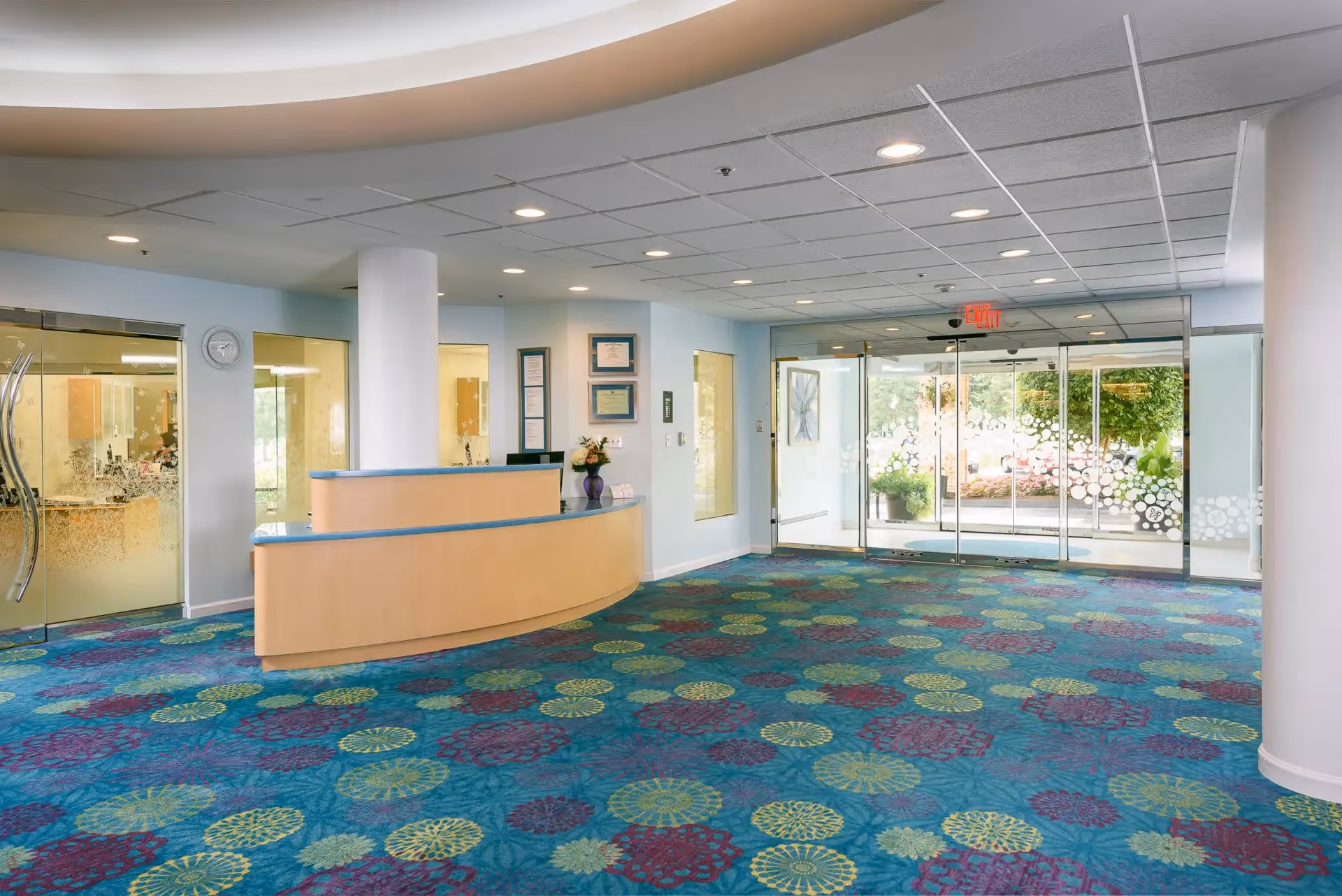 Reception lobby with a curved wooden front desk, colorful patterned blue carpet, and glass automatic entrance doors to the outside.
