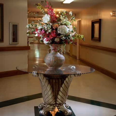 A decorative glass table with an ornate base holding a large vase filled with a colorful arrangement of flowers, placed in the center of a hallway with beige walls and framed artwork.