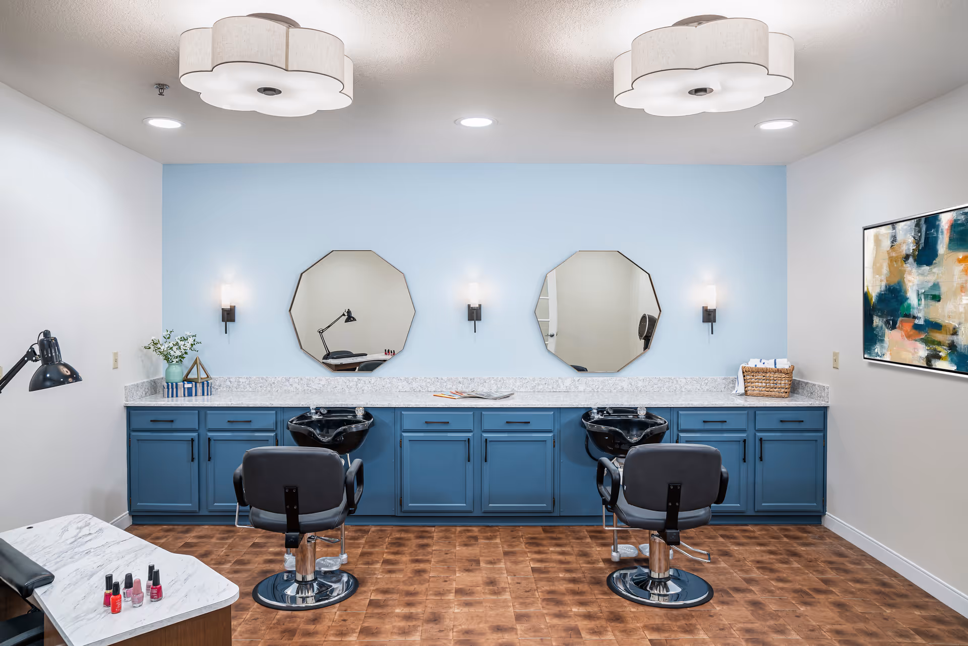 A modern salon area with two black salon chairs in front of a long blue cabinet with granite countertop. Above the countertop are two octagonal mirrors and wall-mounted lights. The room has a light blue accent wall, wooden flooring, and two white ceiling lights shaped like flowers. A small table with nail polish bottles and a lamp is visible on the left side, and a colorful abstract painting hangs on the right wall.