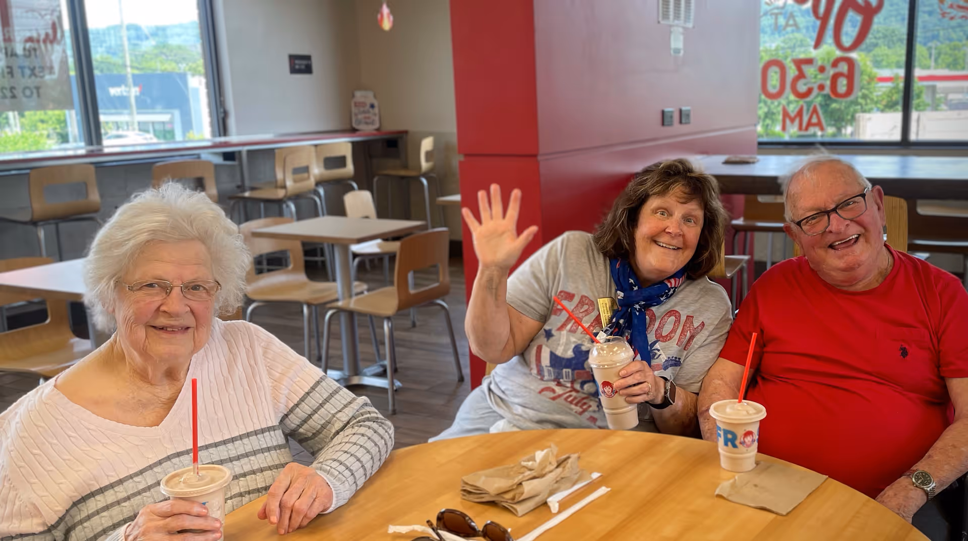Three elderly individuals sitting around a wooden table in a casual dining area. Two women and one man are smiling at the camera, with one woman waving. They each have a drink with a red straw. The background shows empty tables and chairs, large windows, and a red pillar.