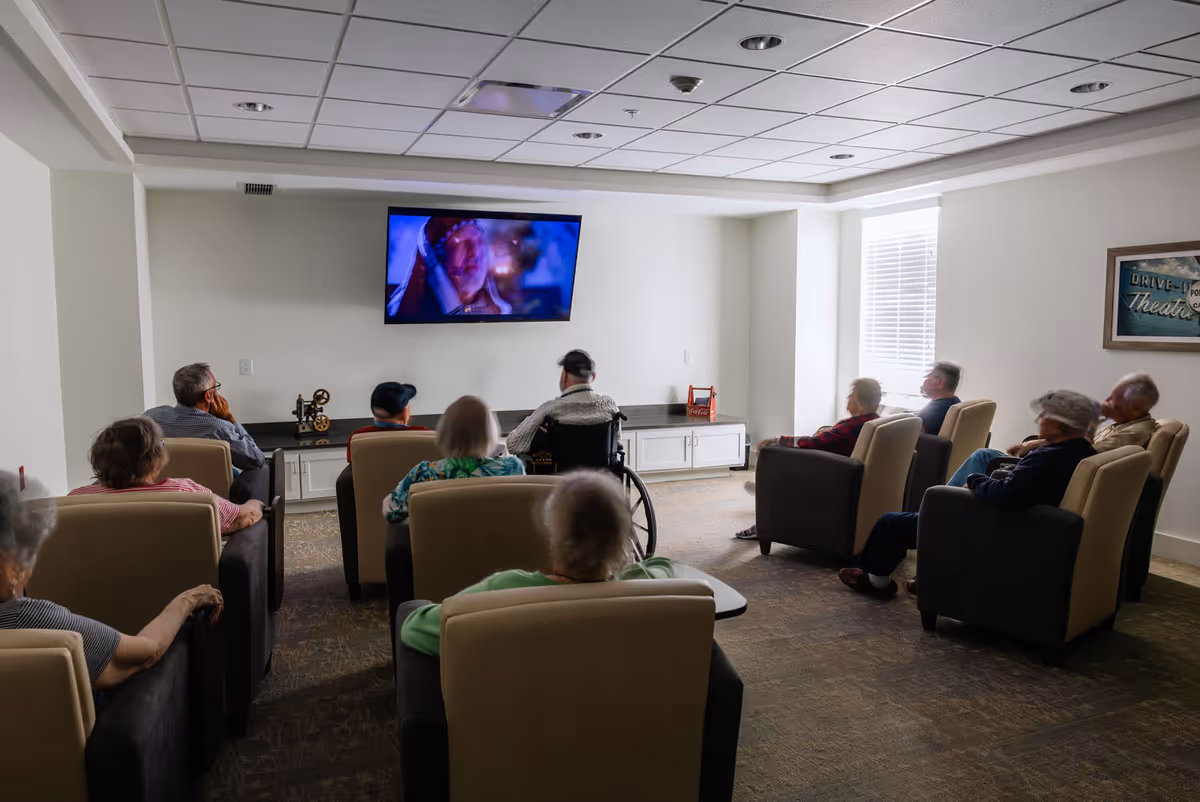 A group of elderly people seated in comfortable armchairs and a wheelchair watching a movie on a wall-mounted TV in a well-lit room with a carpeted floor and a framed picture on the wall.
