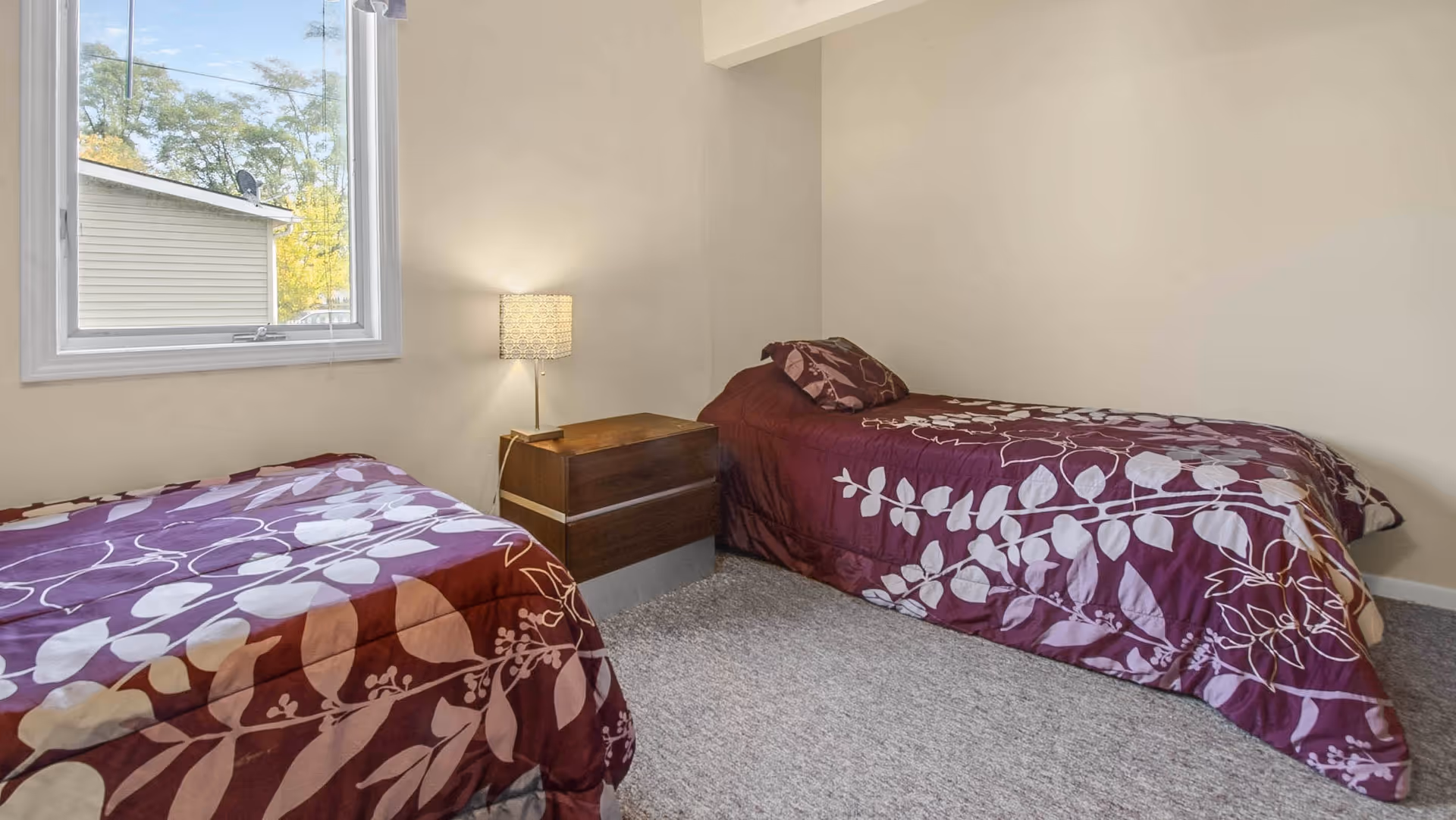 Two twin beds with matching purple floral bedspreads in a simple bedroom with a nightstand and lamp by a window.