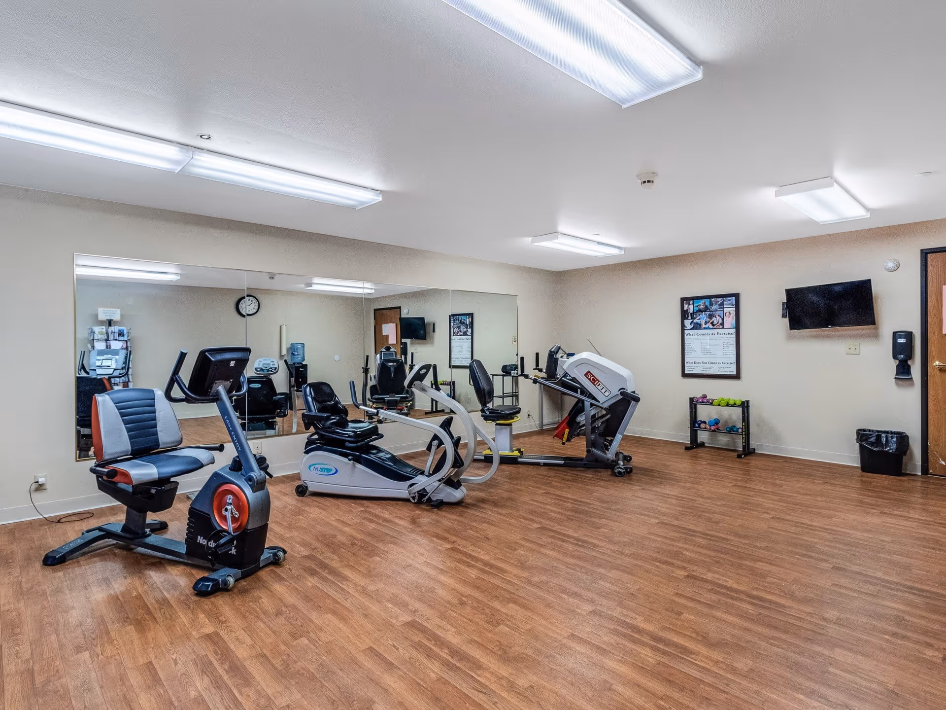 A senior living facility exercise room with wooden flooring, several pieces of exercise equipment including a recumbent bike, a rowing machine, and a leg press machine. The room has large mirrors on one wall, fluorescent ceiling lights, a wall-mounted TV, a small rack with dumbbells, a clock, and a water dispenser.