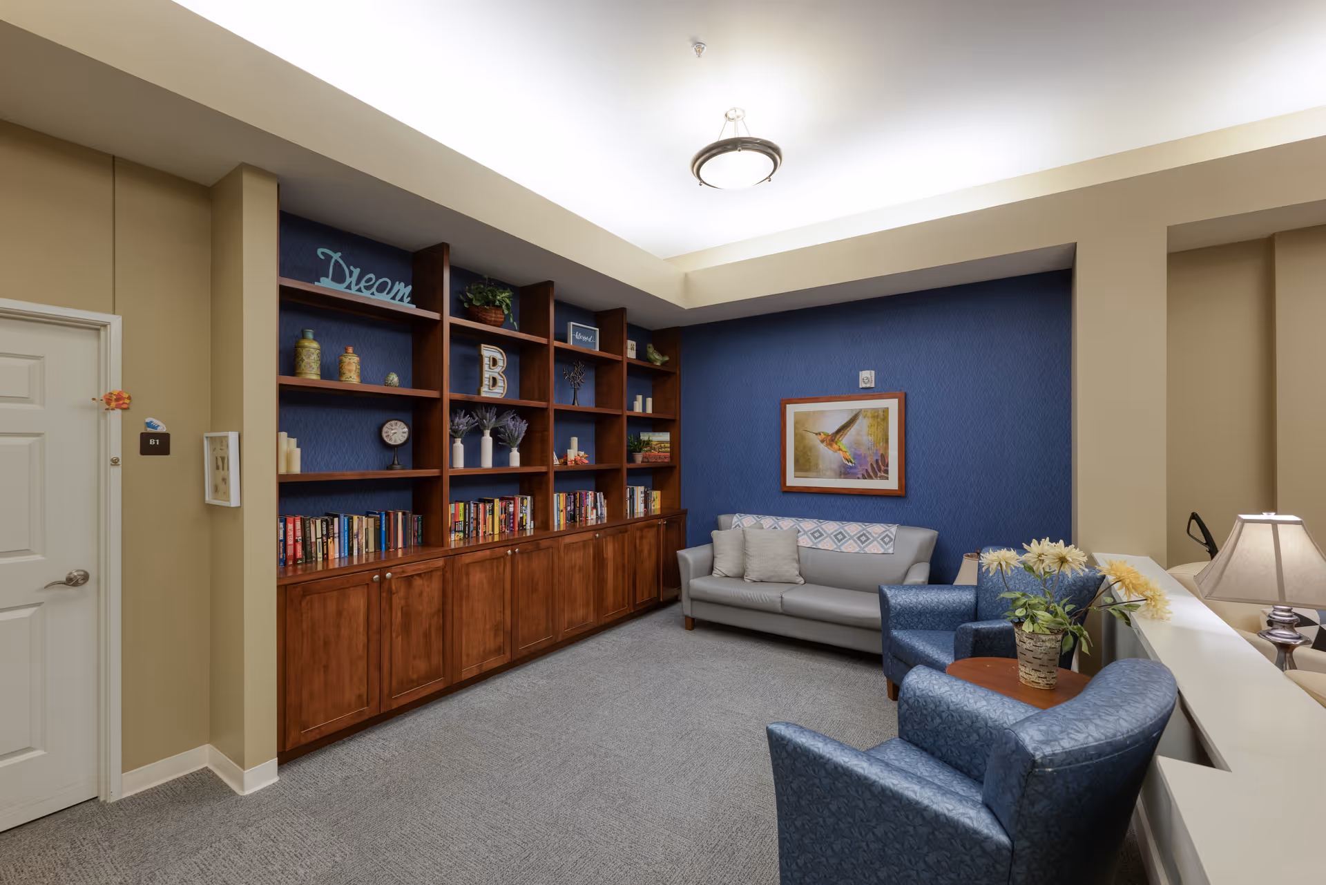 A cozy sitting area in a senior living facility with a gray sofa, two blue armchairs, a wooden bookshelf filled with books and decorative items, a framed hummingbird painting on a blue accent wall, and a small table with a flower arrangement. The room has beige walls, gray carpet, and soft overhead lighting.