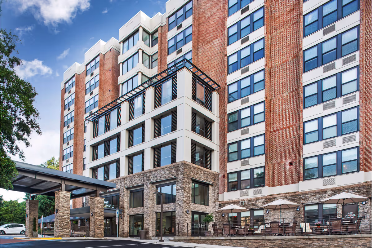 Exterior view of a multi-story senior living facility with brick and stone facade, large windows, a covered entrance, and outdoor seating area with tables and umbrellas.