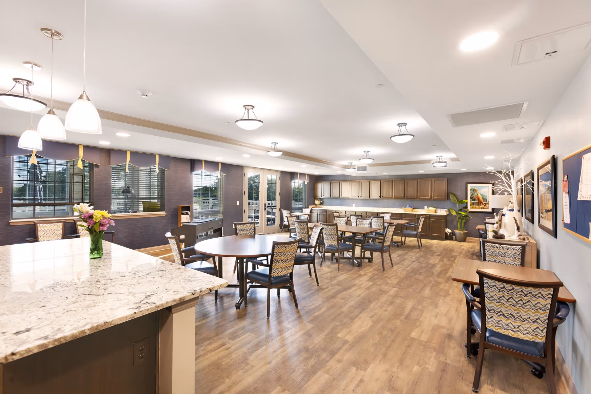 A spacious and well-lit dining room with multiple round and rectangular tables surrounded by chairs. The room features large windows with blinds, a granite countertop with pendant lights above, wooden flooring, and cabinets along the far wall. There are decorative elements including a vase with flowers on the countertop and framed artwork on the walls.
